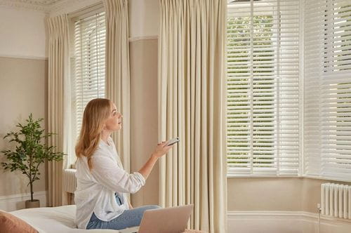 Une femme tient une télécommande et la pointe vers des stores automatisés. Elle est assise sur un lit dans une chambre lumineuse et élégamment meublée, avec des rideaux crème et une plante en pot.