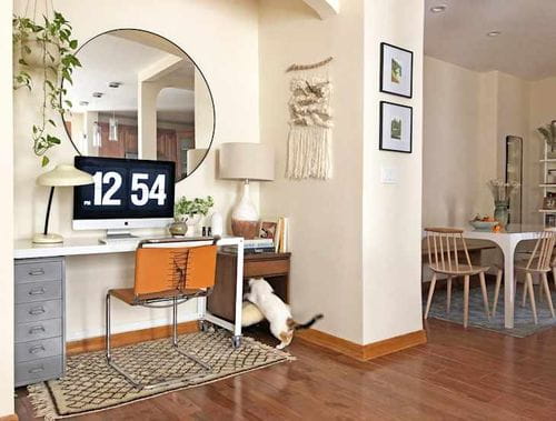 Bureau à domicile avec un bureau, un ordinateur, un grand miroir rond et un chat qui explore une étagère. Coin repas visible en arrière-plan.