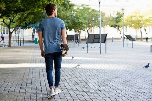 Jeune homme marchant sur un trottoir avec un skateboard - photos et images libres de droits d'un homme marchant de dos
